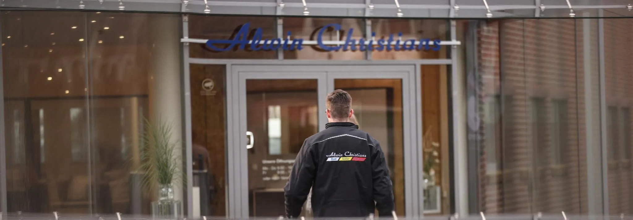 Ein Anlagenmechaniker mit Firmenjacke von Alwin Christians steht mit dem Rücken zur Kamera und nähert sich einem modernen Büro- oder Geschäftseingang mit Glasfront.