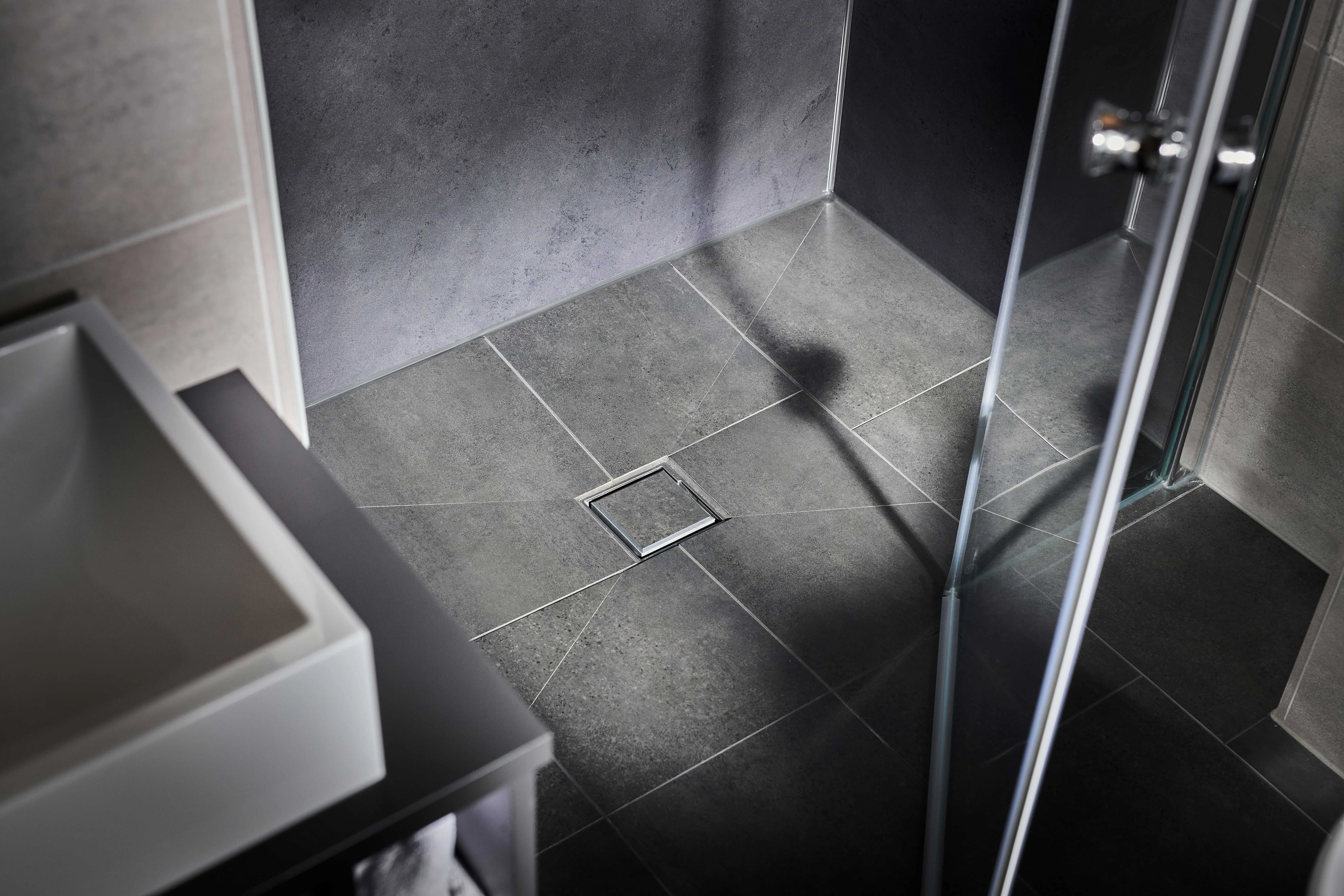 Detailaufnahme einer bodengleichen, gefliesten Dusche mit mittigem quadratischem Bodenablauf und dunkelgrauen Wandflächen.