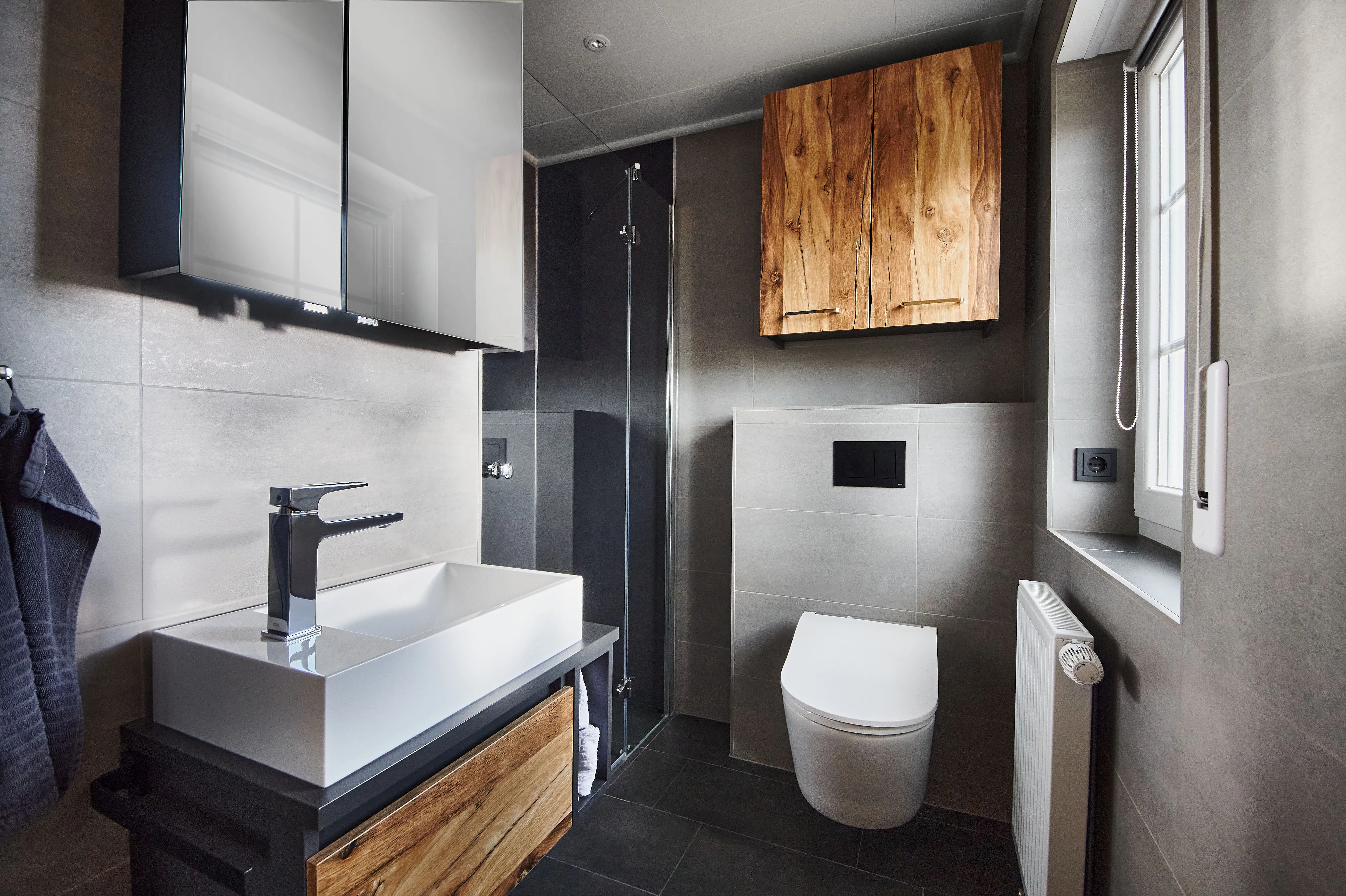 Modernes Bad mit wandhängendem WC, ebenerdiger Glasdusche, großem weißen Aufsatzwaschbecken und Holzmöbeln in warmer Maserung.