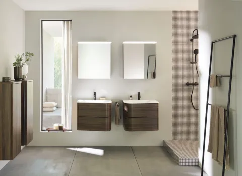 Stilvolles Bad mit hängenden Holz-Badezimmermöbeln der Serie badu von burgbad sowie Doppelwaschbecken und beleuchteten Spiegeln. Eine bodenebene Dusche mit Mosaikfliesen ergänzt das Design.