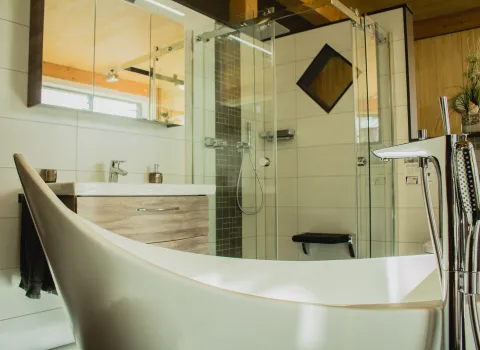 Modernes Badezimmer mit freistehender Badewanne, Glasdusche, Holzoptik-Unterschrank und hellen Fliesen in warmer Atmosphäre.