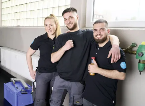 Drei junge Auszubildende im SHK-Handwerk posieren lächelnd in Arbeitskleidung vor einer Werkstattwand mit großem Fenster.