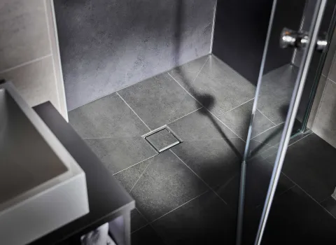 Detailaufnahme einer bodengleichen, gefliesten Dusche mit mittigem quadratischem Bodenablauf und dunkelgrauen Wandflächen.