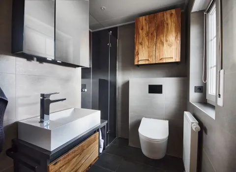 Modernes Bad mit wandhängendem WC, ebenerdiger Glasdusche, großem weißen Aufsatzwaschbecken und Holzmöbeln in warmer Maserung.