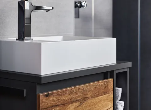 Nahaufnahme eines modernen Waschtischs mit weißem Aufsatzbecken, schwarz-matter Armatur und Front in natürlicher Holzoptik.
