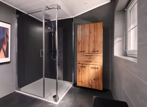 Großer Duschbereich mit Glasabtrennung, dunkler Wandverkleidung und Duschwanne; daneben ein Hochschrank in markanter Holzoptik sowie ein Fenster mit breiter Laibung.