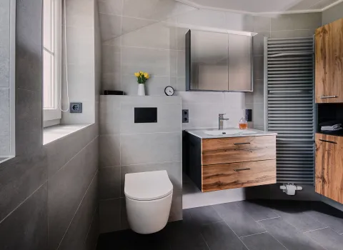 Modernes Badezimmer im Obergeschoss mit wandhängendem WC, Waschtischmöbel in Holzoptik, großem Spiegelschrank, grauen Feinsteinzeugfliesen und seitlichem Designheizkörper.