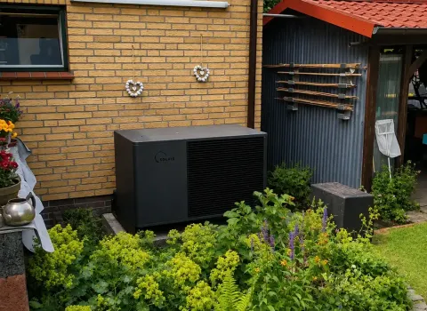 Eine kompakte Wärmepumpen-Außeneinheit ist inmitten eines grünen Hausgartens aufgestellt und wird teilweise von der umliegenden Bepflanzung eingerahmt. Das dunkle, kubische Gerät befindet sich in unmittelbarer Nähe zur Backsteinfassade des Gebäudes. Die Gartensituation veranschaulicht, wie sich moderne Wärmepumpentechnik unauffällig in einen bestehenden Außenbereich integrieren lässt.