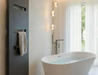 Freistehende Badewanne vor einem großen Fenster mit Blick ins Gruene. Links ein flacher Designheizkoerper in Dunkelgrau mit einem haengenden Handtuch.