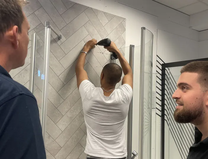 Ein Mann bohrt mit einer Bohrmaschine in eine Wand mit hellen Fliesen im Fischgrätmuster, während zwei Kollegen ihn während des Techniktrainings beobachten.