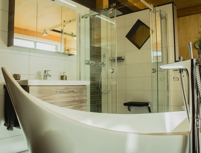 Modernes Badezimmer mit freistehender Badewanne, Glasdusche, Holzoptik-Unterschrank und hellen Fliesen in warmer Atmosphäre.