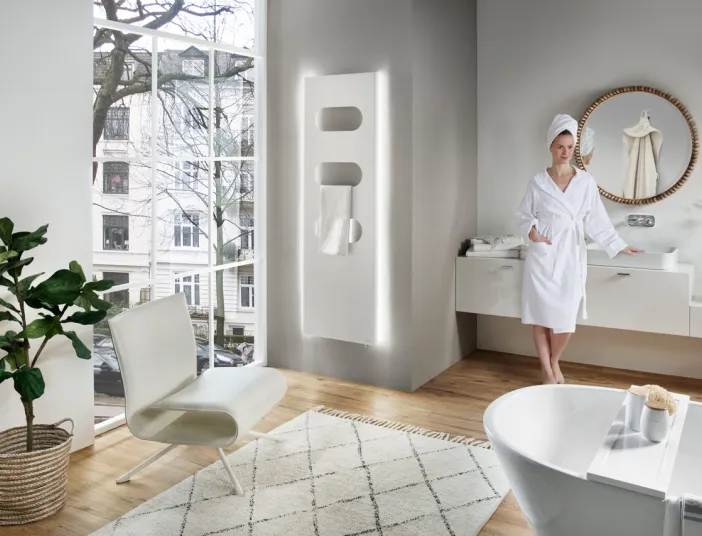 Eine Frau in einem weißen Bademantel und mit einem Handtuch um den Kopf steht in einem modernen Badezimmer.